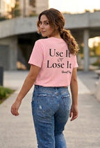 A woman wearing a light pink graphic t-shirt with the text 'Use It or Lose It' and 'Unsettled Thing' printed in black, paired with blue jeans, walking outdoors on a concrete path.