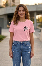 A woman wearing a light pink crewneck t-shirt with 'Go you!' printed in black script on the left chest, paired with distressed blue jeans, standing outdoors on a paved walkway.
